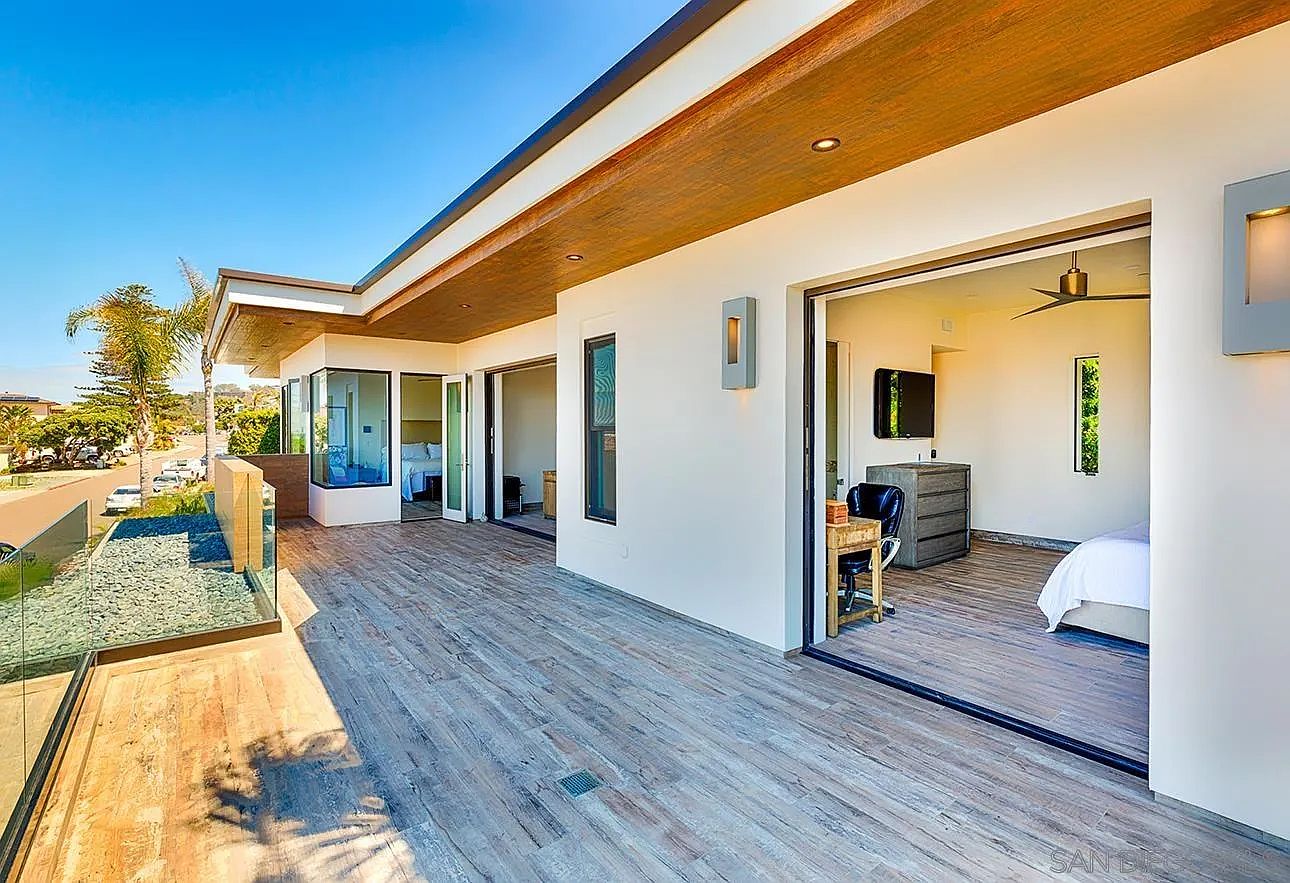 This image showcases a spacious outdoor patio or balcony area, featuring wood-look tile flooring and glass railings that offer unobstructed views of the surrounding landscape. The area connects seamlessly to the interior through large sliding glass doors, creating an indoor-outdoor living experience. The design incorporates modern architectural elements, such as recessed lighting and clean lines, enhancing the property's appeal.