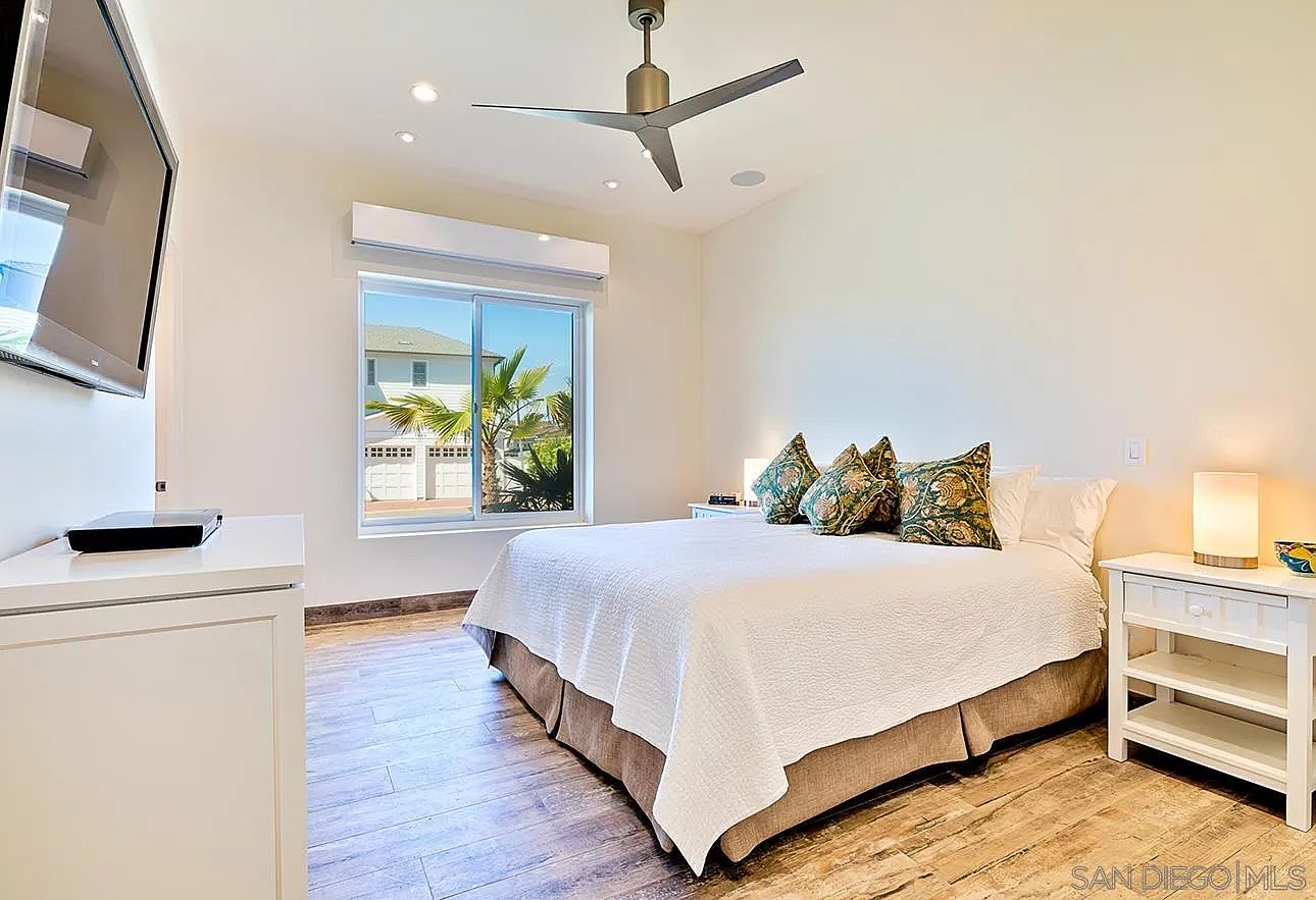 This is a bright and airy primary bedroom featuring a queen-sized bed with white linens and decorative pillows. The room has hardwood floors, a ceiling fan, and a large window offering natural light. A white nightstand with a lamp sits beside the bed, and a dresser with a TV is on the opposite wall.