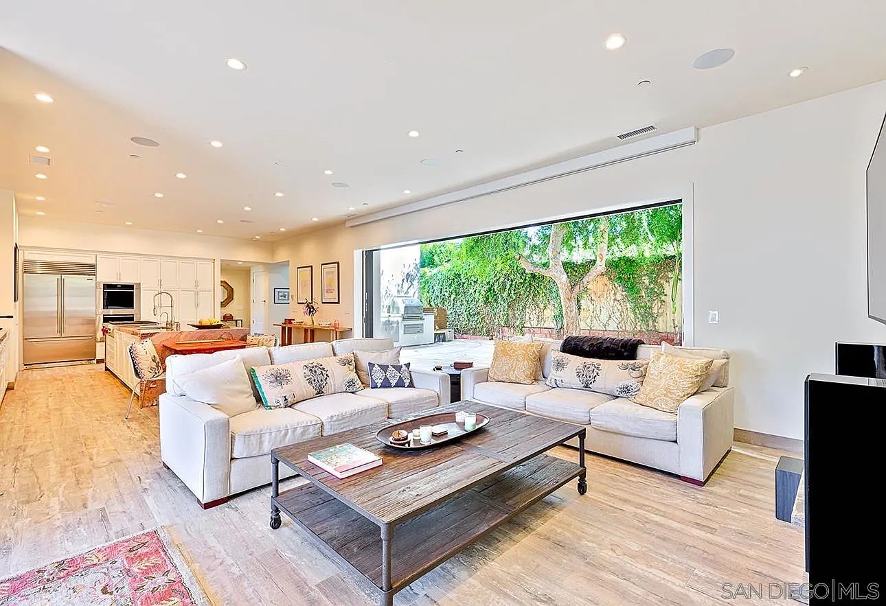 This is an interior shot of a living room that seamlessly blends indoor and outdoor living. The room features two large, comfortable sofas arranged around a rustic coffee table, with a wide opening leading to a lush backyard. The flooring is light wood, and the overall aesthetic is bright, airy, and inviting, perfect for relaxation and entertaining.