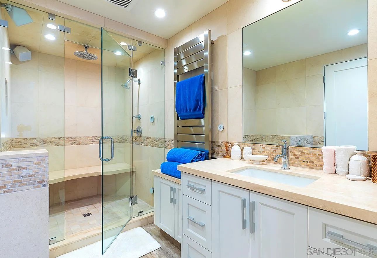 This is a well-lit primary bathroom featuring a glass-enclosed shower with a built-in bench and a rainfall showerhead. The vanity has white cabinets, a light-colored countertop, and a modern sink and faucet. A heated towel rack and a large mirror add to the luxurious feel of the space.