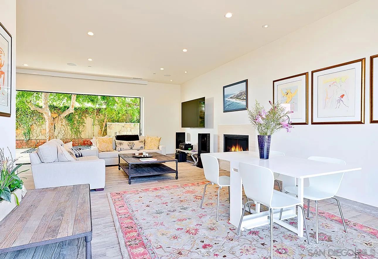 This is an interior shot of a living and dining area. The living room features a large window overlooking a lush garden, a comfortable sofa, and a modern coffee table. The dining area includes a white table with four chairs, complemented by artwork on the wall and a stylish rug that ties the spaces together, creating a bright and inviting atmosphere.