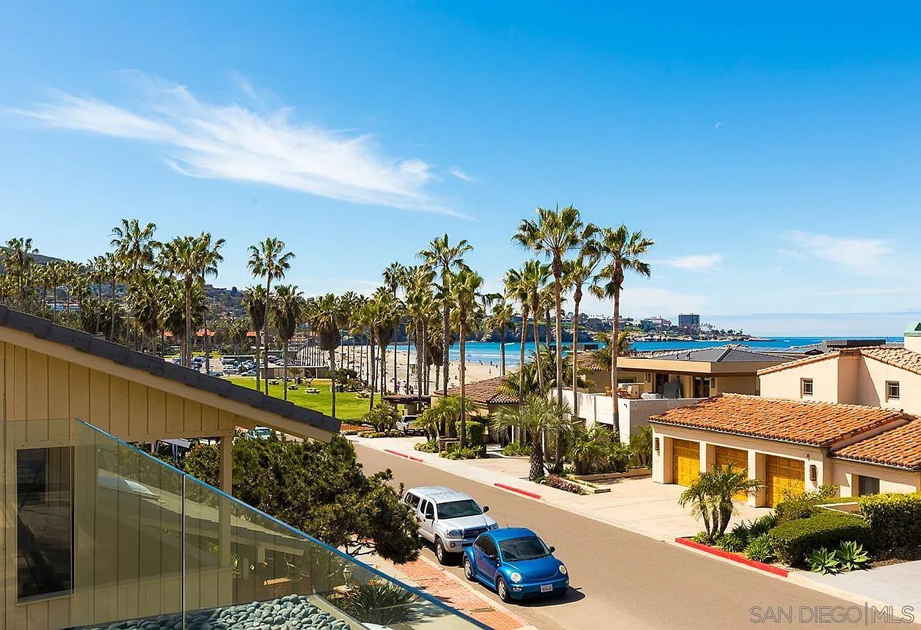 This aerial view showcases a coastal property with a clear blue sky backdrop. Palm trees line the street leading to a sandy beach, and well-maintained homes with terracotta roofs are visible. Two cars are parked on the street, adding a touch of everyday life to the scene.