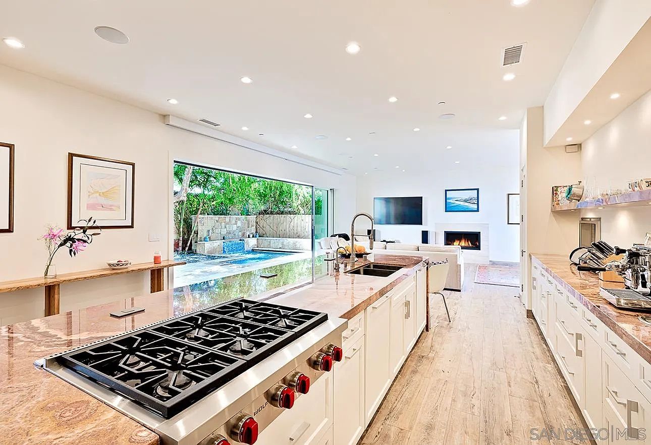 This is a bright and spacious kitchen featuring a large island with a cooktop and marble countertops. The kitchen has white cabinetry and stainless steel appliances, with an open layout that flows into the living area. A large sliding glass door provides access to the outdoor pool area, creating a seamless indoor-outdoor living experience.