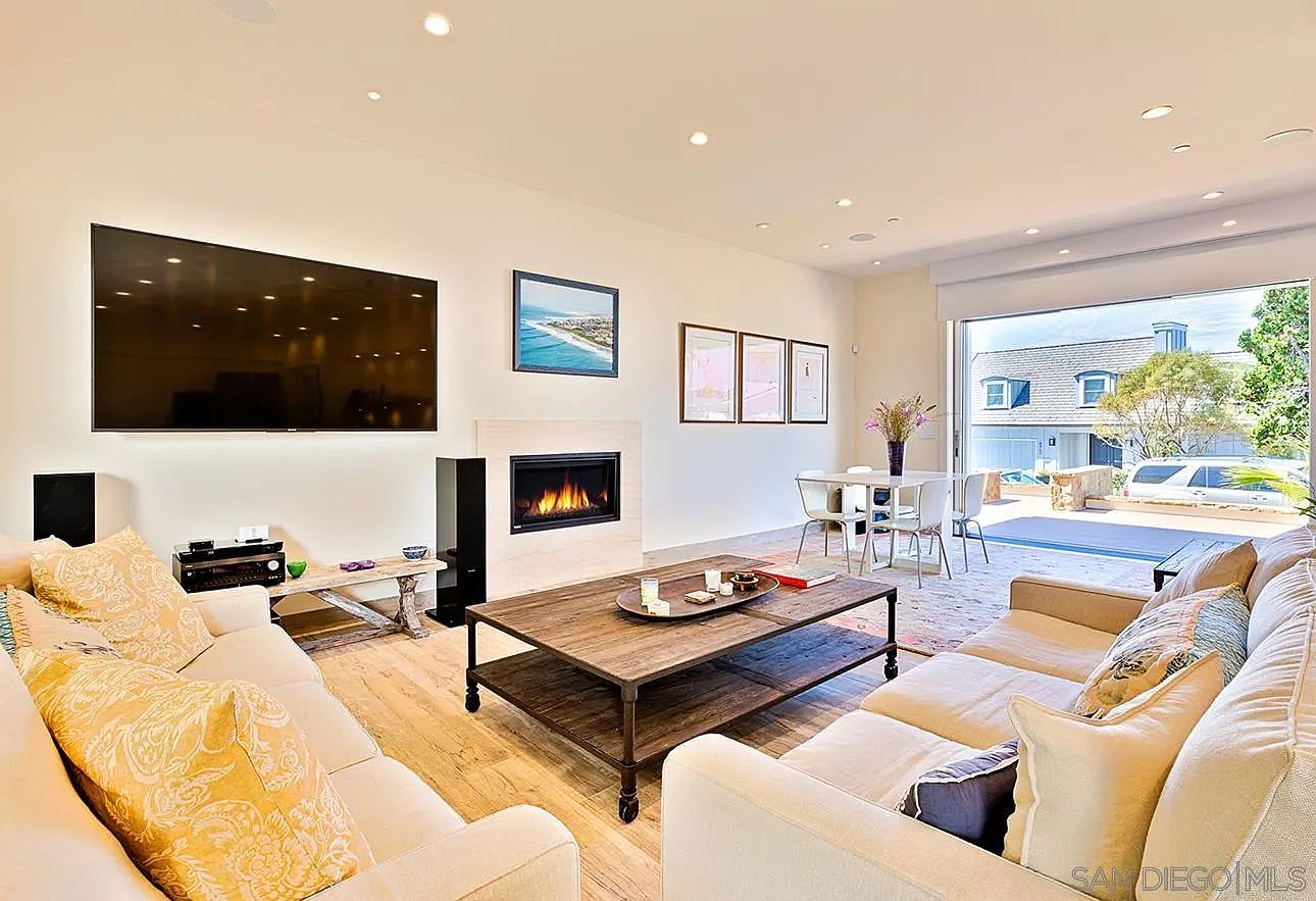 This is a bright and spacious living room featuring hardwood floors, a modern fireplace, and comfortable seating arrangements. A large television is mounted on the wall, and natural light floods the room through a large sliding glass door that opens to the outside. The overall impression is one of comfort and contemporary style.