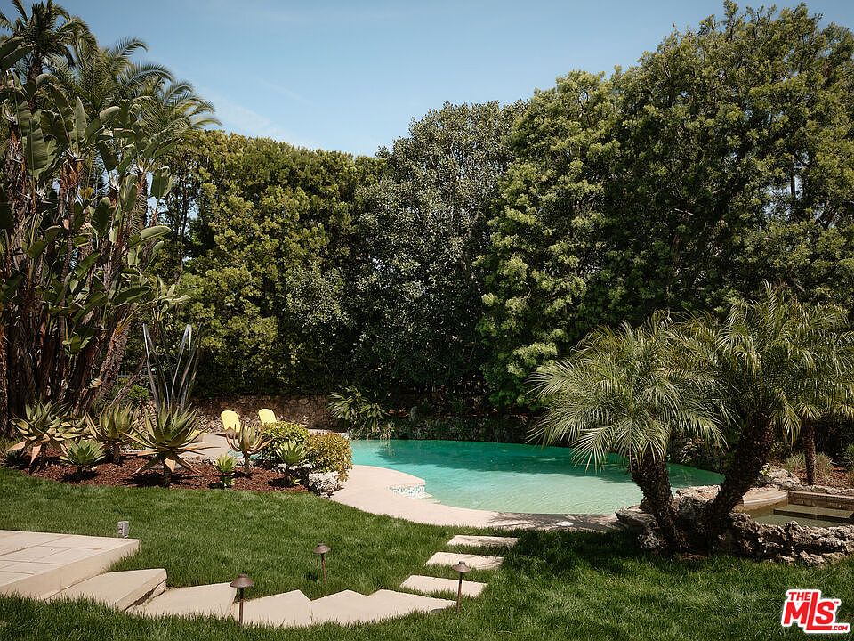 This image showcases a luxurious backyard pool area, surrounded by lush greenery and mature trees, creating a private and serene oasis. Stone steps lead to the pool, which features clear turquoise water, and the landscaping includes a variety of plants and palm trees, enhancing the tropical ambiance. The scene evokes a sense of relaxation and upscale outdoor living.