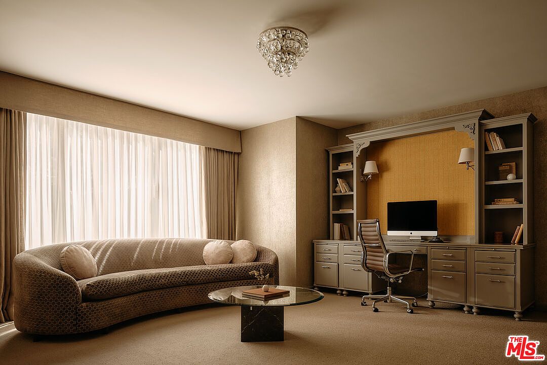 This interior shot showcases a well-appointed office or study. A curved sofa sits near a window with sheer curtains, while a built-in desk and shelving unit with a computer and chair occupy the opposite side of the room. A chandelier hangs from the ceiling, adding a touch of elegance to the space.