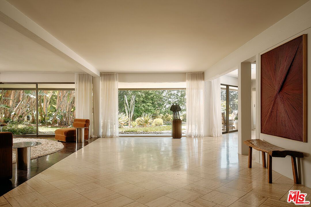 This is a spacious living room featuring large windows that offer views of the lush garden outside. The room has a neutral color palette with travertine tile flooring and sheer white curtains that diffuse natural light. A modern art piece hangs on the wall, complemented by a rustic wooden bench, adding a touch of sophistication and warmth to the space.