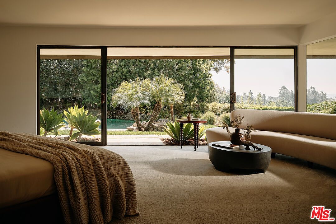 This primary bedroom features a seamless indoor-outdoor transition with large sliding glass doors opening to a lush garden. The room is furnished with a low-profile bed and a curved sofa, complemented by a round coffee table. The neutral color palette and natural light create a serene and inviting atmosphere.