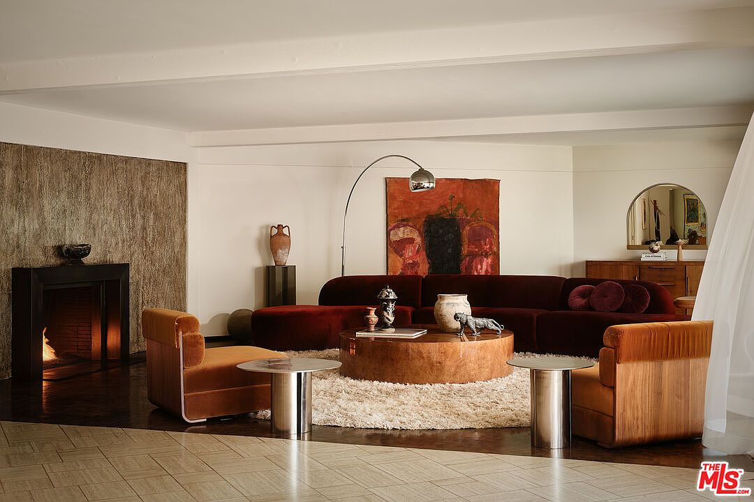 This is a stylish living room featuring a large, curved, burgundy sectional sofa, a round wooden coffee table on a shag rug, and two orange armchairs flanking the seating area. A modern arc lamp illuminates the space, and artwork adds a touch of sophistication. The room has a warm and inviting atmosphere, perfect for relaxation and entertaining.