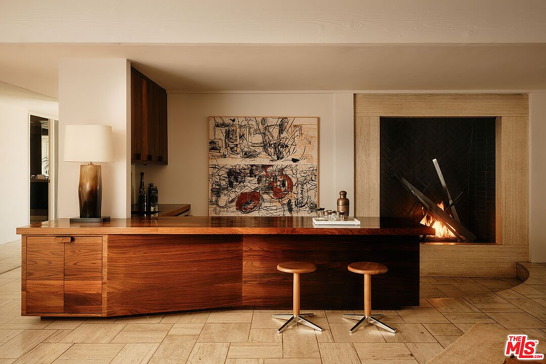 This interior shot showcases a modern living room with a custom wooden bar area, featuring sleek cabinetry and two bar stools. An abstract art piece hangs above the bar, and a contemporary fireplace with a stone surround adds warmth and visual interest. The room is finished with neutral-toned tile flooring, creating a sophisticated and inviting atmosphere.