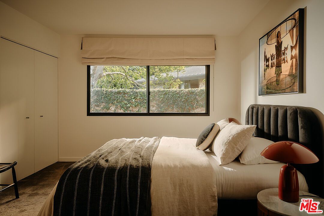 This is a cozy primary bedroom featuring a large window with a beige roman shade, a dark upholstered headboard, and neutral-toned bedding. A modern art piece hangs above the bed, and a unique red lamp sits on a round side table. The room has a calm and inviting atmosphere.
