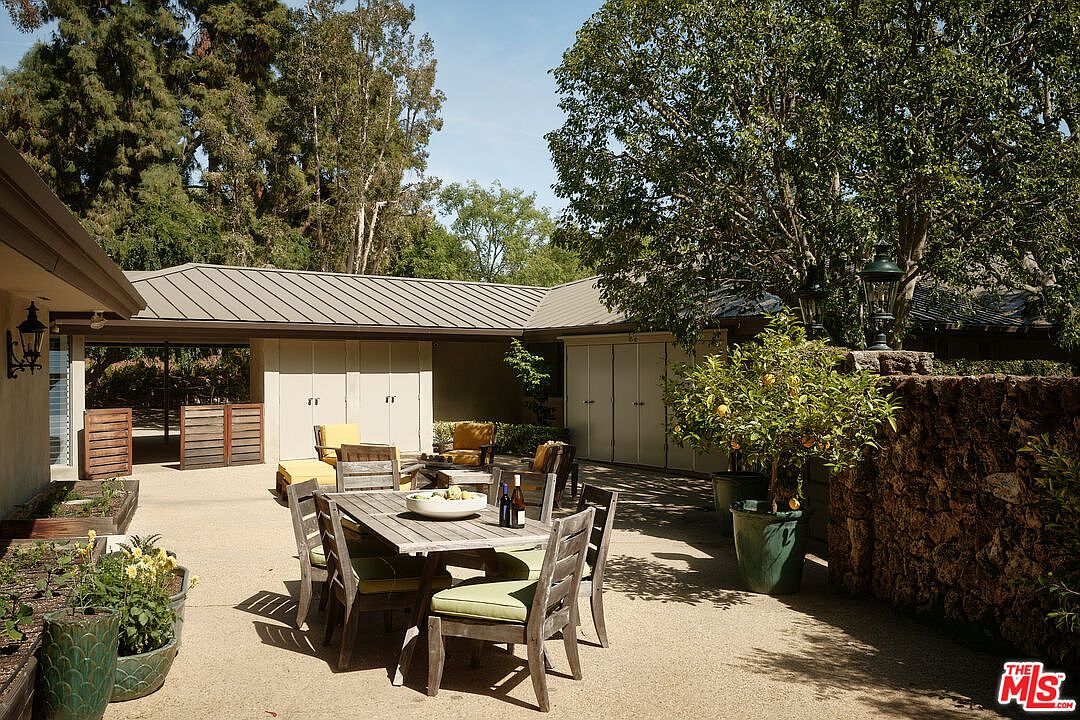 This image showcases an inviting outdoor patio area, perfect for entertaining or relaxing. A wooden dining table with chairs sits prominently in the center, complemented by comfortable lounge seating in the background. The patio is surrounded by lush greenery, including potted plants and mature trees, creating a serene and private atmosphere.