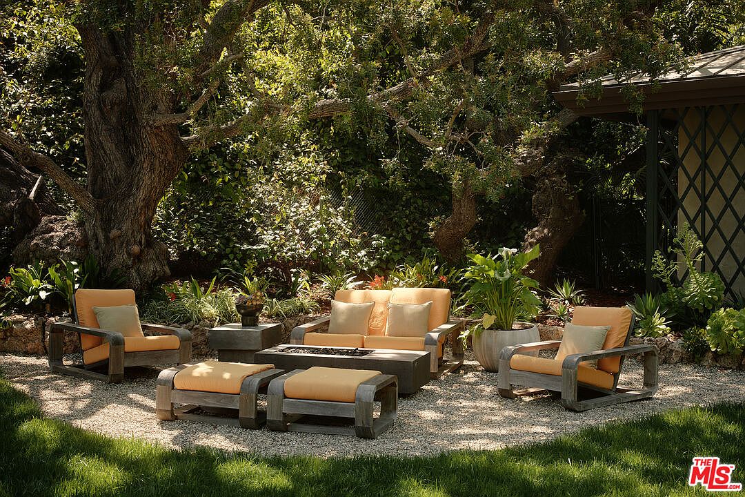 This image showcases a beautifully arranged outdoor seating area, featuring a matching set of chairs, a loveseat, ottomans, and a coffee table with a fire pit. The furniture is crafted from wood with comfortable cushions, set on a gravel surface surrounded by lush greenery and mature trees, creating a serene and inviting outdoor living space.
