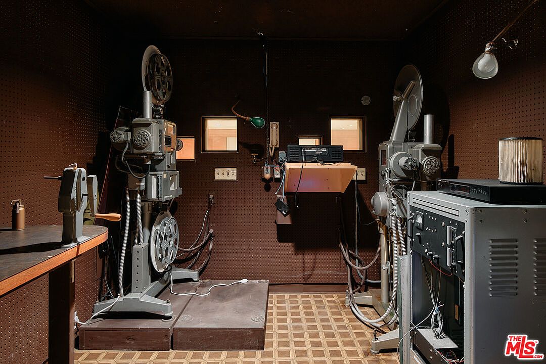 This is an interior shot of a vintage film projection room. The room features two large film projectors, one on each side, along with a desk and various electrical components. The walls are covered in a dark, textured material, and the floor has a patterned tile, creating a retro and functional space.