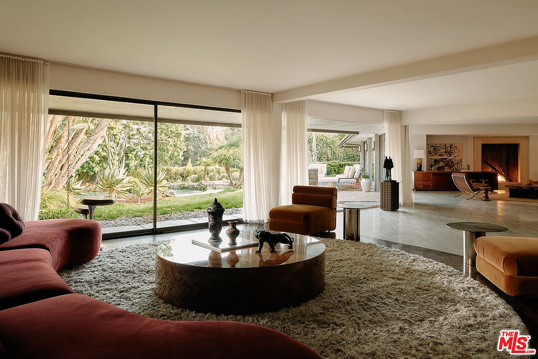 This is an interior shot of a luxurious living room featuring a curved, maroon-colored sofa and a round, textured coffee table with decorative objects. Large windows offer a view of a lush garden, and the open floor plan leads to an adjacent space with additional seating. The room exudes a modern and sophisticated ambiance.