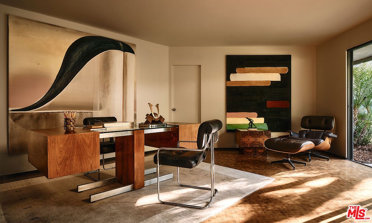 This interior shot showcases a stylish office space featuring a wooden desk with chrome accents, complemented by a black leather chair. Artwork adorns the walls, and a comfortable lounge chair with an ottoman sits in the corner, creating a sophisticated and inviting atmosphere. The room is well-lit, highlighting the parquet flooring and the overall mid-century modern design.