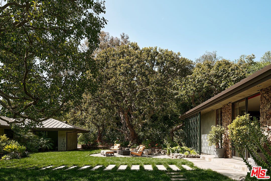 This image showcases a beautifully landscaped yard with a modern architectural style home in the background. The yard features lush green grass, stone pathways, and mature trees providing ample shade. A cozy outdoor seating area with a fire pit adds a touch of luxury and relaxation, making it an inviting space for entertaining or unwinding.