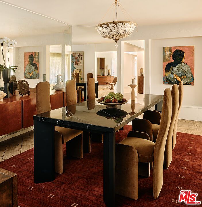 This is an interior shot of a dining room featuring a large, dark marble-topped table with black legs, surrounded by tall, upholstered chairs in a warm brown tone. A modern chandelier hangs above the table, and artwork adorns the walls, creating a sophisticated and inviting atmosphere. The perspective is from a medium distance, showcasing the entire dining set and its placement within the room.