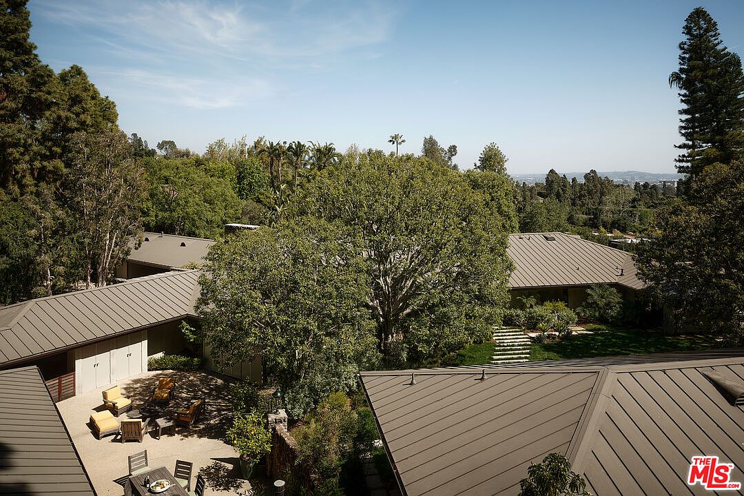This aerial view showcases a sprawling estate nestled among lush greenery. The property features multiple structures with sleek, modern roofs, connected by landscaped pathways and courtyards. The overall impression is one of privacy, luxury, and seamless integration with the natural surroundings.