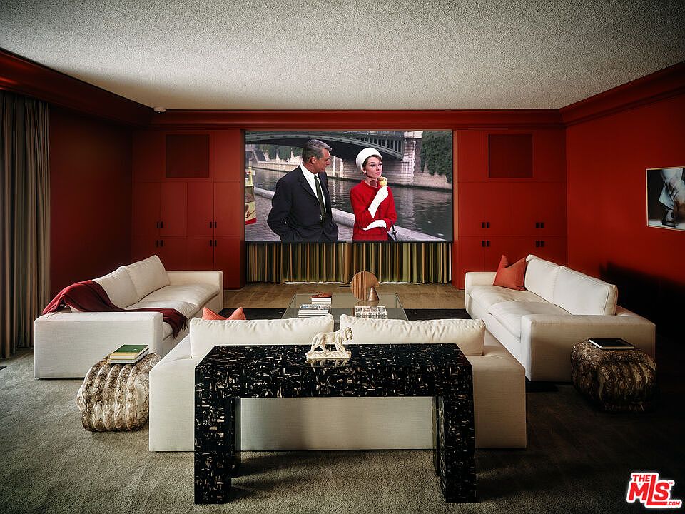 This is an interior shot of a living room featuring a large screen displaying a scene from a movie. The room is decorated in a red color scheme with white sofas arranged around a central coffee table and a decorative console table. The perspective is from the center of the room, showcasing the symmetrical arrangement of the furniture and the focal point of the screen.