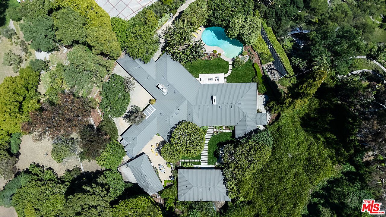This aerial view showcases a sprawling estate nestled among lush greenery. The property features a large, multi-winged house with a gray roof, a swimming pool, and meticulously landscaped grounds. The overall impression is one of luxury, privacy, and expansive outdoor living.