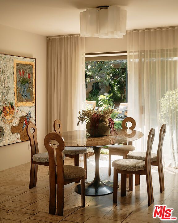 This interior shot showcases a dining room with a round table and unique wooden chairs. Natural light filters through sheer curtains, illuminating the room and highlighting the artwork on the wall. The overall ambiance is elegant and inviting, perfect for intimate gatherings.