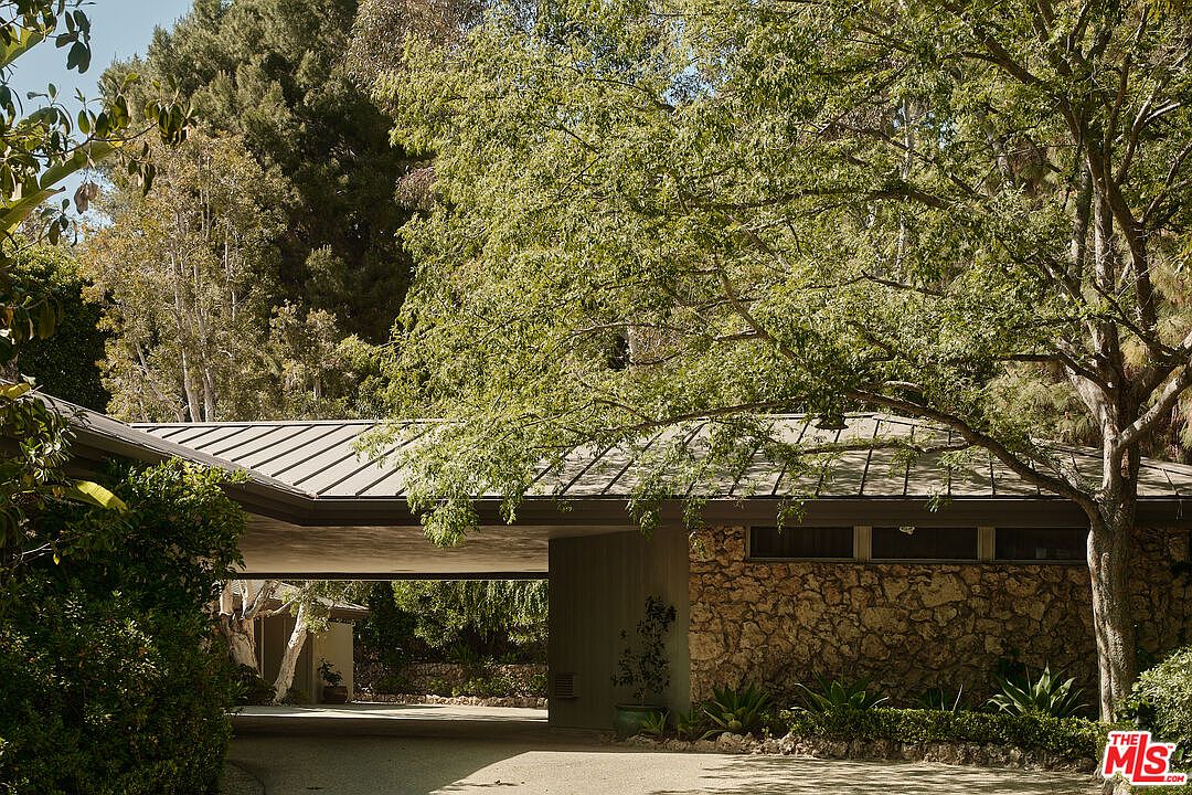 The image showcases the front exterior of a mid-century modern home, characterized by its low-slung roofline, stone facade, and integrated carport. Lush greenery surrounds the property, creating a sense of privacy and natural beauty. The architectural style emphasizes horizontal lines and a seamless integration with the landscape.