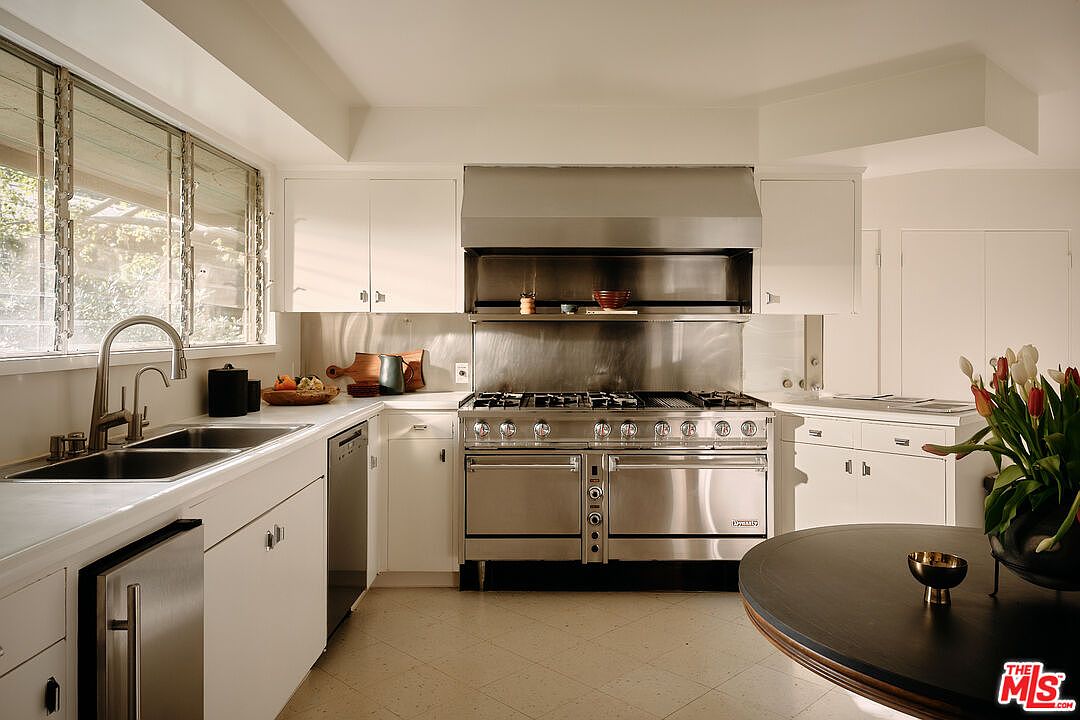 This is a well-lit kitchen featuring white cabinets, stainless steel appliances, and a light-colored tile floor. A large stainless steel range hood and stove are the focal point, while a sink and countertop area are visible to the left. The kitchen has a clean and functional design, with a round table adding a touch of warmth.