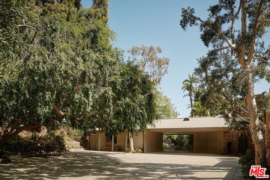 The image showcases the front exterior of a modern home, emphasizing its unique architectural design. A covered carport connects two sections of the house, framed by mature trees that provide shade and privacy. The driveway is spacious, and the overall aesthetic blends natural elements with contemporary construction.