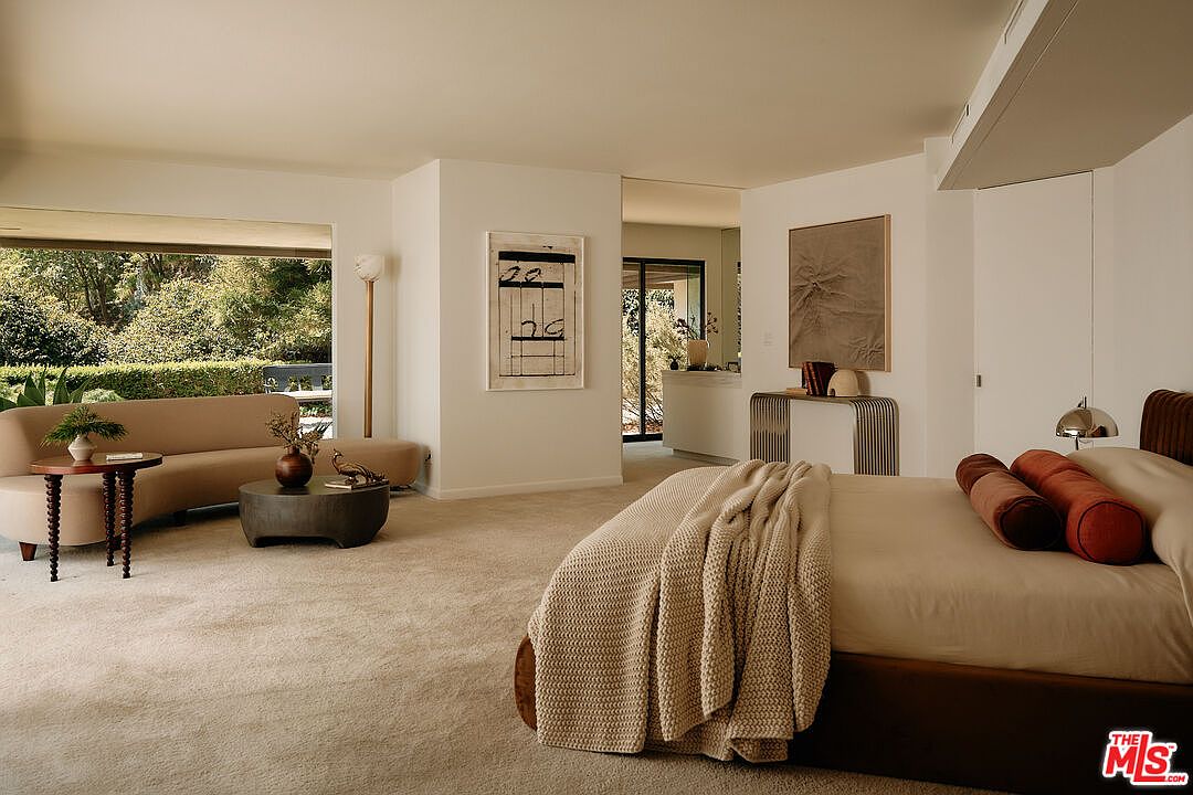 This is a bright and airy primary bedroom featuring a large bed with neutral linens and decorative pillows. The room includes a seating area with a curved sofa, round coffee table, and side table, creating a comfortable and inviting space. Large windows offer views of the lush greenery outside, enhancing the room's tranquil atmosphere.