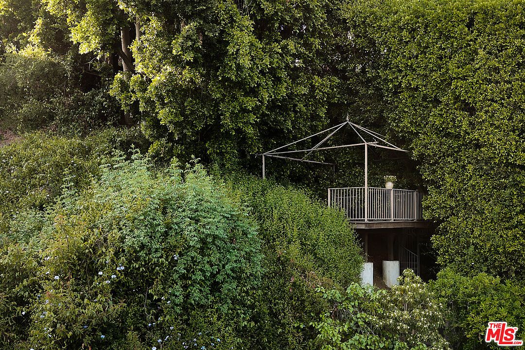This image showcases a lush, overgrown garden featuring a charming gazebo-like structure nestled among the greenery. The structure has a metal frame and railing, adding a touch of architectural interest to the natural setting. The overall impression is one of a secluded and tranquil outdoor space, perfect for relaxation and enjoying nature.