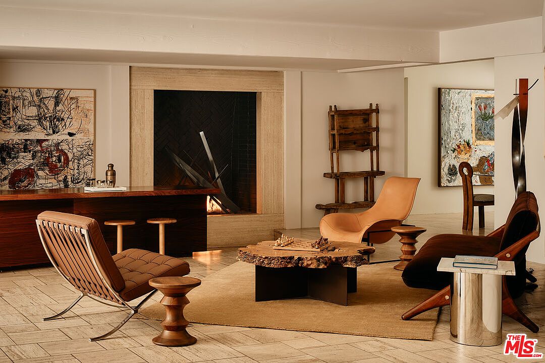 This interior shot showcases a stylish living room with a focus on unique furniture and art. The room features a modern fireplace, a wooden desk with bar stools, and several distinctive chairs arranged around a rustic coffee table with a chess set. The overall aesthetic is sophisticated and artistic, creating a warm and inviting atmosphere.