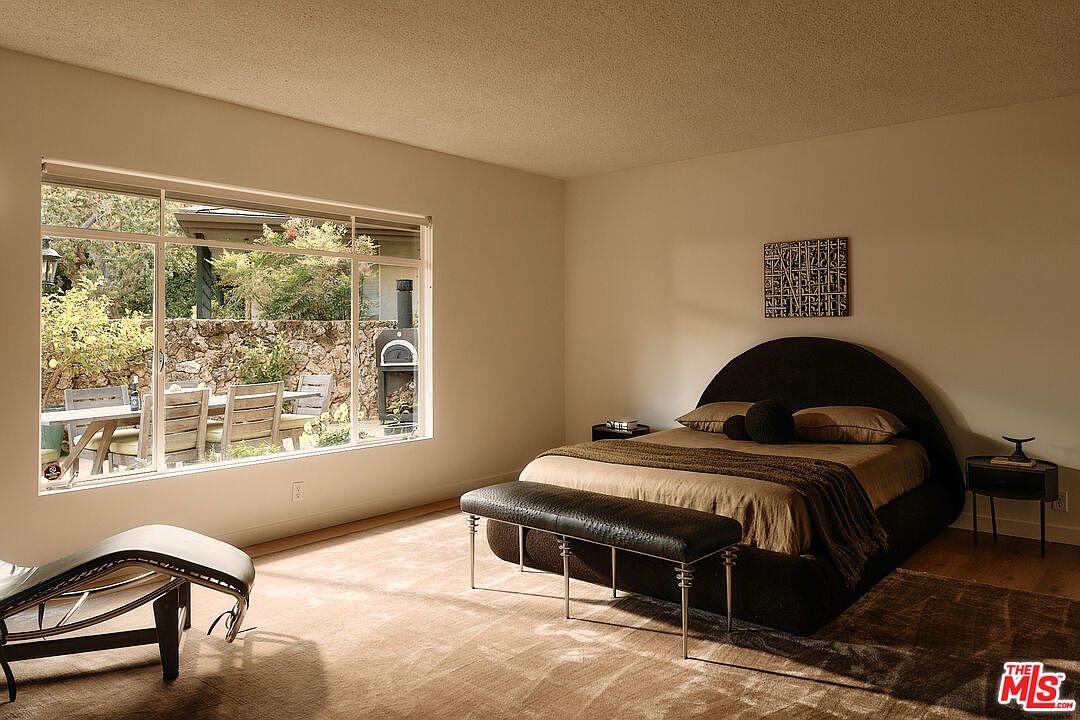 This is a bedroom featuring a large window with a view of an outdoor dining area and stone wall. The room is furnished with a unique, rounded headboard bed, a dark leather bench, and a modern side table. The color palette is neutral, creating a calm and inviting atmosphere.