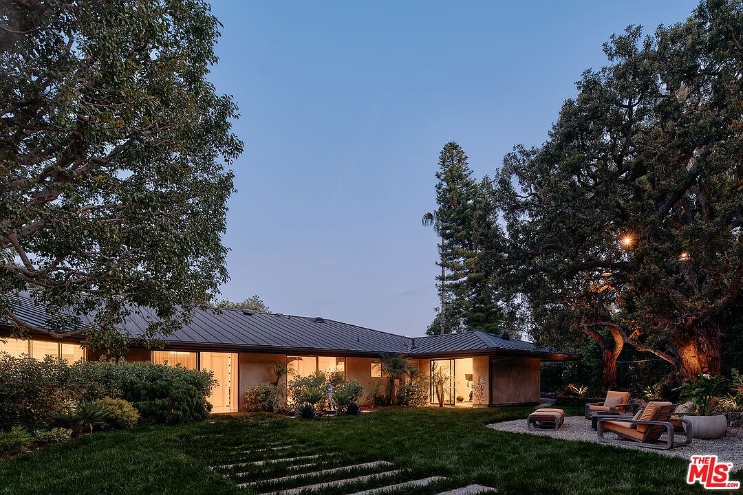 This image showcases the rear exterior of a modern home at dusk, featuring a dark metal roof and expansive glass windows that illuminate the interior. The well-manicured lawn is accented with stone pathways, leading to a cozy outdoor seating area with stylish chairs and a small table, all nestled beneath mature trees with string lights, creating an inviting and serene atmosphere.