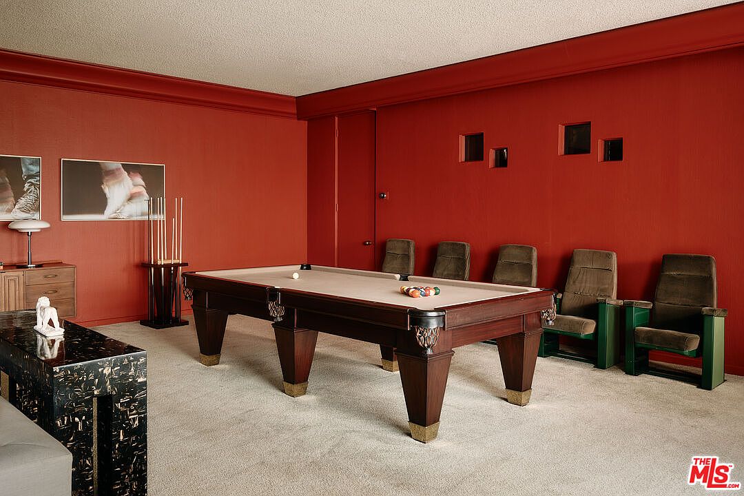 This interior shot showcases a game room featuring a pool table as the central element, surrounded by red walls and unique design choices. The room includes several chairs along one wall, artwork, and a distinctive black and gold table, creating a sophisticated yet playful atmosphere. The carpet is light-colored, providing a contrast to the darker tones of the furniture and walls.