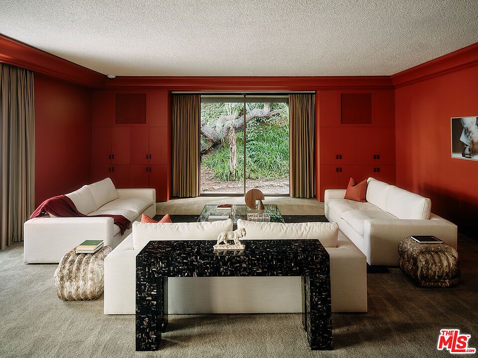 This living room features a symmetrical layout with two white sofas facing each other, flanking a glass coffee table. A black console table sits in the foreground, adding a modern touch. The room is painted in a deep red hue, creating a warm and inviting atmosphere, while large windows offer a view of the greenery outside.