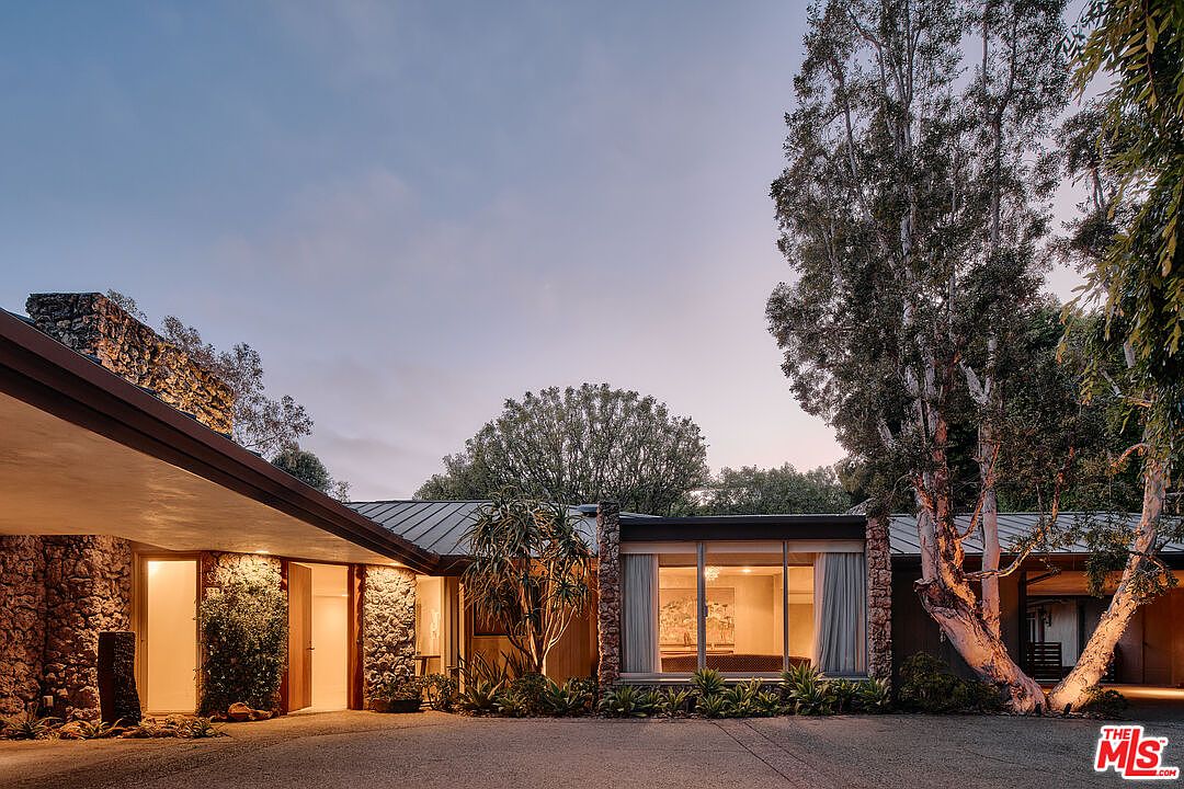 The image showcases the front exterior of a mid-century modern home at dusk. The house features a low-slung roofline, stone facade elements, and large windows that offer a glimpse into the warmly lit interior. Mature trees and landscaping surround the property, adding to its secluded and inviting atmosphere.