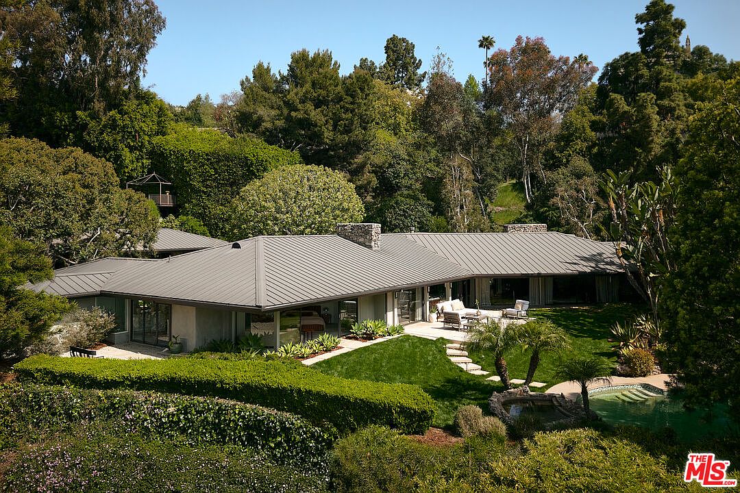 This aerial view showcases a sprawling single-story home with a modern design, featuring a low-pitched, gray metal roof and large windows. The property is surrounded by lush greenery, including mature trees, manicured hedges, and a well-maintained lawn. A swimming pool is visible in the backyard, adding to the property's appeal.