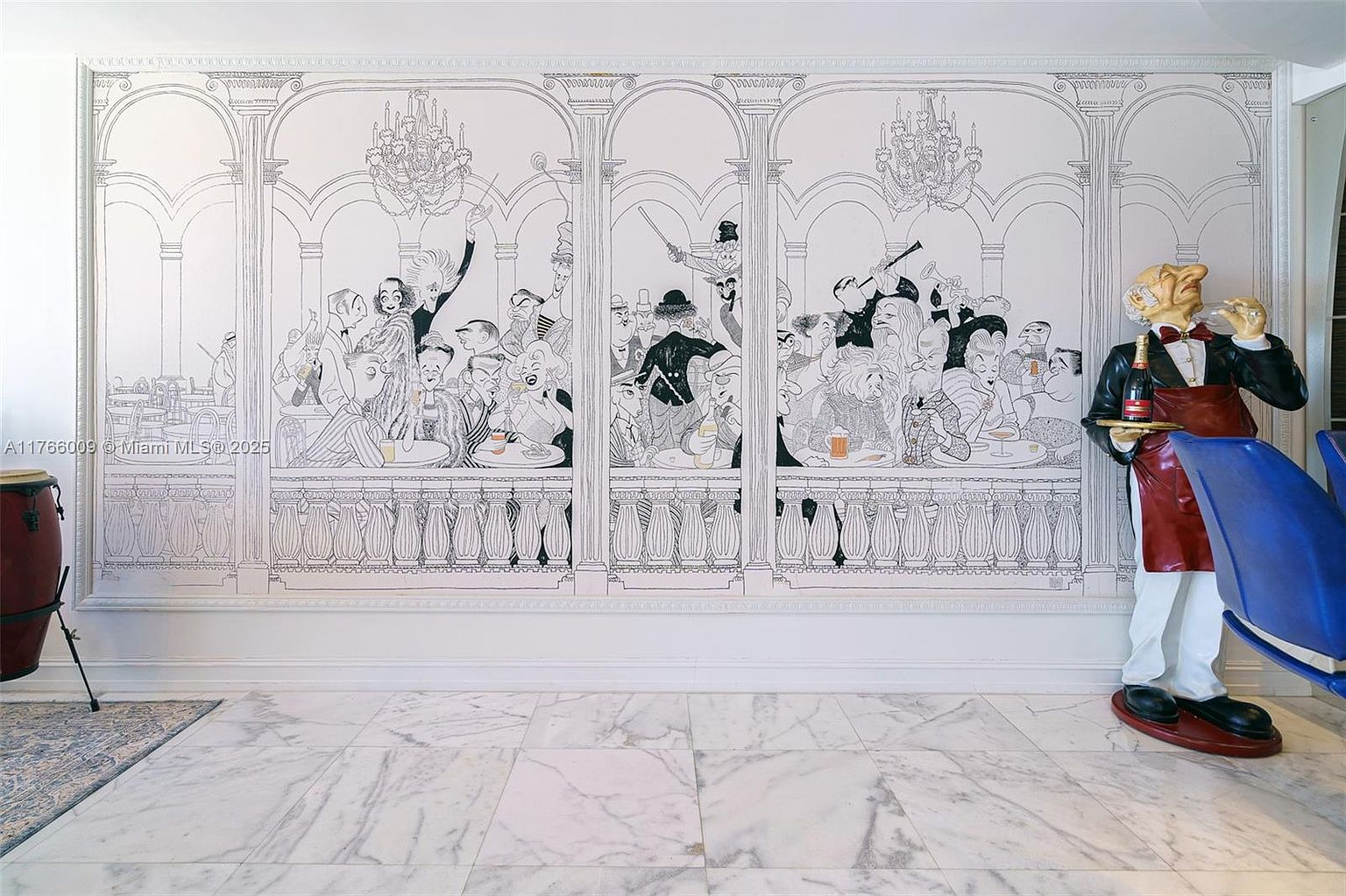 This interior space features a prominent, large-scale mural depicting a lively, illustrated scene of people dining in a grand hall. To the right, a whimsical, life-sized waiter statue holds a tray with a bottle of champagne, adding a playful, artistic touch to the room. The floor is finished with light-colored, polished stone tiles, and the overall aesthetic is eclectic and highly stylized.