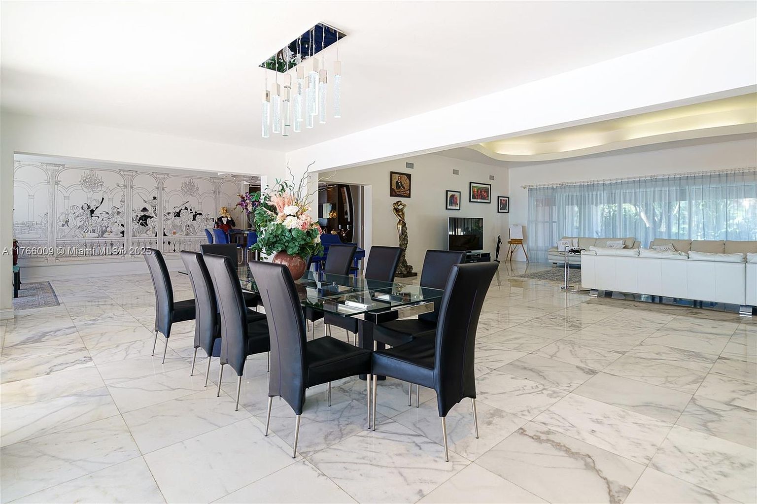 This spacious, open-concept dining area features a glass-topped table surrounded by six high-backed black chairs, set against a backdrop of a large, intricate mural. The room is finished with polished light-colored stone flooring and a modern, multi-pendant light fixture hanging above the table. To the right, the space flows into a bright living area with white sofas and large windows, creating an expansive and elegant atmosphere.