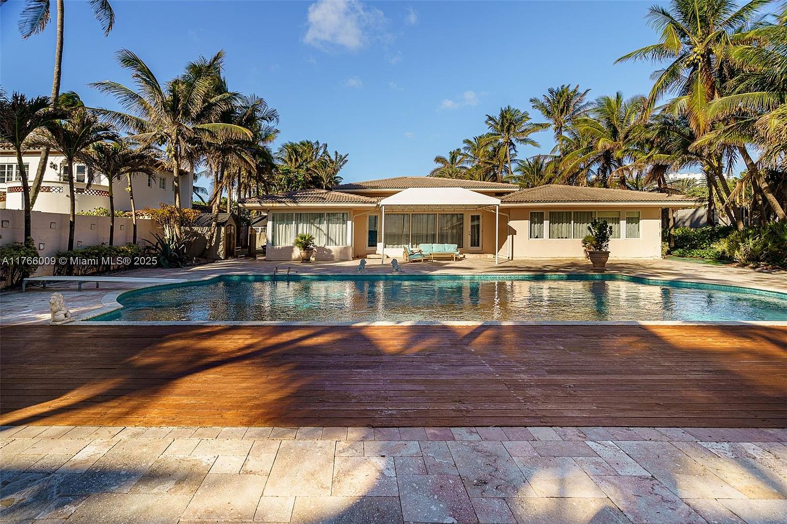 This rear exterior view showcases a spacious swimming pool set against a single-story residence surrounded by lush palm trees. A large wooden deck transitions into a stone-paved patio area, providing ample space for outdoor lounging under a covered patio structure. The scene captures a serene, tropical atmosphere with clear blue skies and well-maintained landscaping.