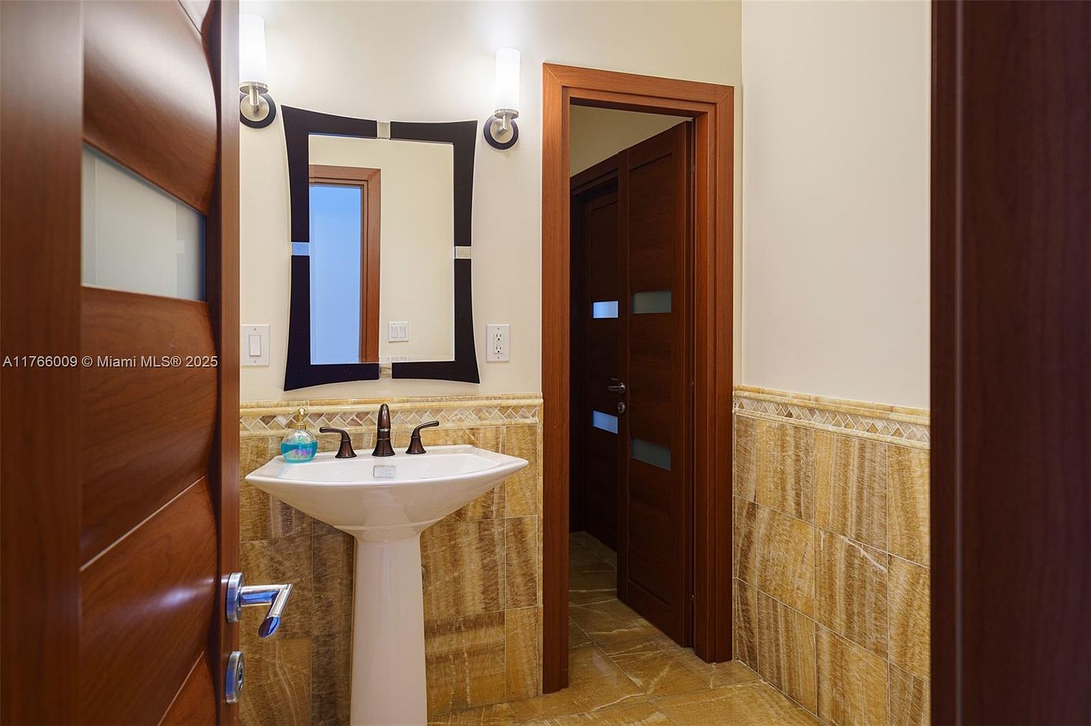 This compact powder room features a pedestal sink with a dark-toned faucet set against a wall of warm-toned, textured stone tiles. A modern, geometric-framed mirror hangs above the sink, flanked by two wall-mounted light fixtures. The space is framed by rich, dark wood door frames, creating a sophisticated and cohesive aesthetic.