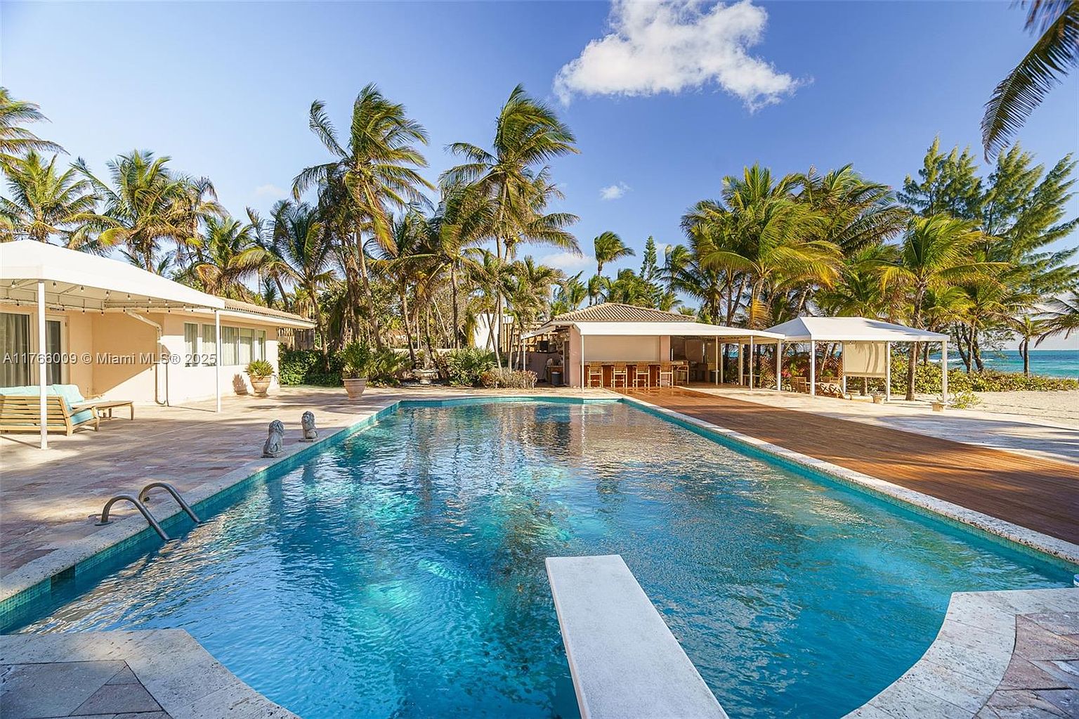 This expansive outdoor living space features a large, rectangular swimming pool with a diving board as the central focal point. The surrounding area includes a stone-paved patio, a wooden deck section, and covered structures providing shaded seating and bar areas. Lush palm trees and tropical landscaping frame the view, which opens up to a sandy beach and the ocean in the distance.