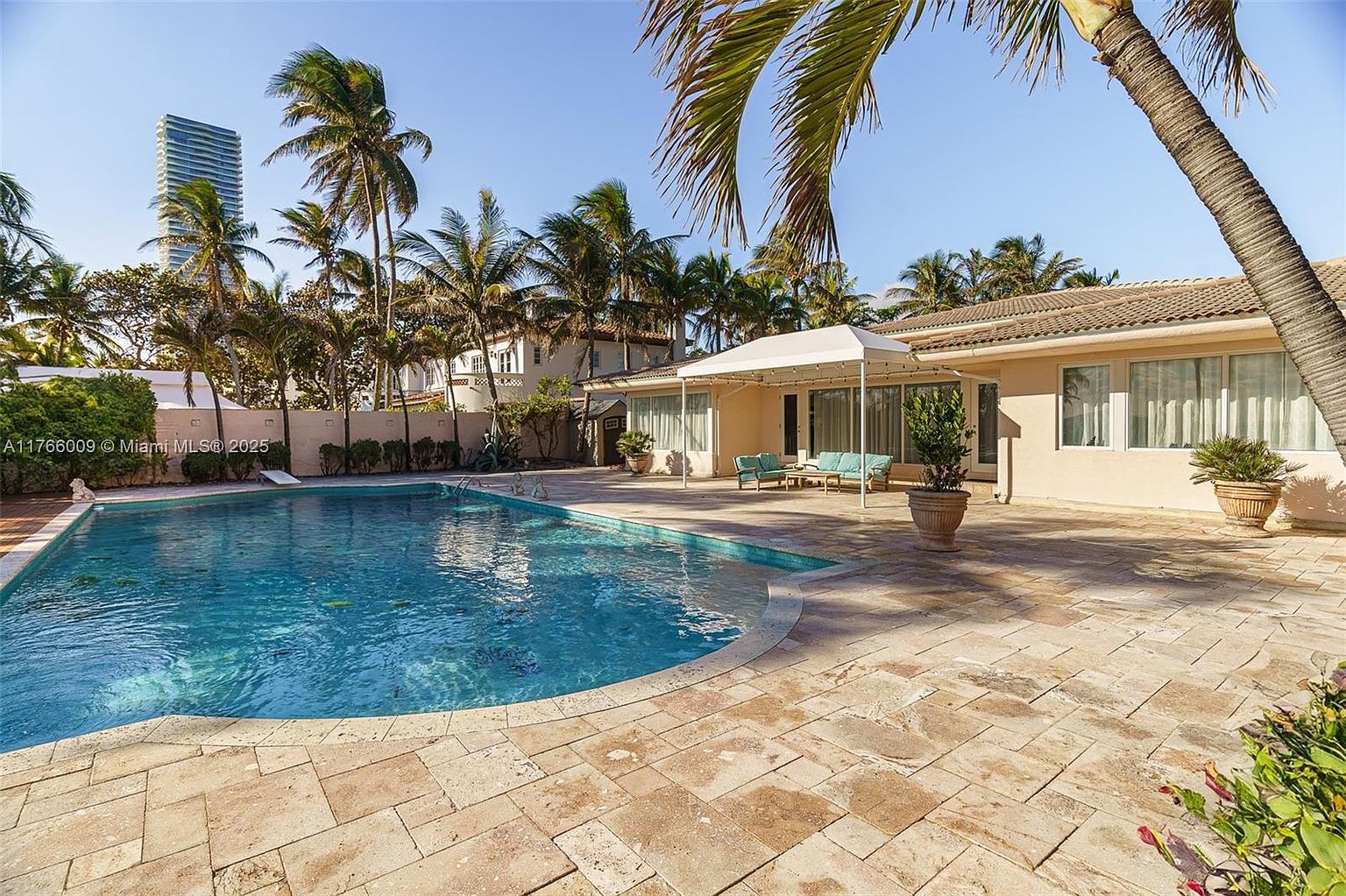 This expansive outdoor living space features a large, free-form swimming pool set against a backdrop of lush palm trees and a clear blue sky. A spacious stone-paved patio surrounds the pool, offering a shaded seating area under a white canopy, perfect for relaxation or entertaining. The scene captures a serene, tropical atmosphere with a view of a neighboring high-rise building in the distance.