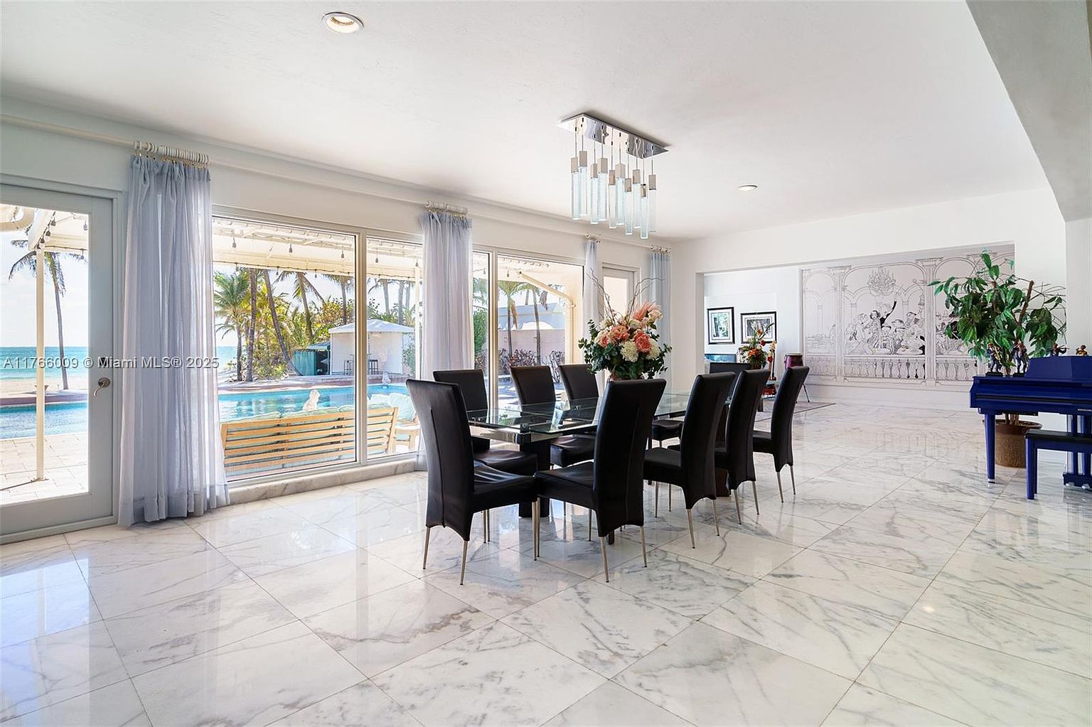 This spacious dining area features a large glass-topped table surrounded by eight black chairs, set against a backdrop of expansive windows that provide a clear view of the outdoor pool and beach. The room is finished with polished light-colored stone flooring and a modern, multi-pendant chandelier hanging above the table. To the right, a vibrant blue piano sits near a decorative wall mural, creating a sophisticated and airy atmosphere.