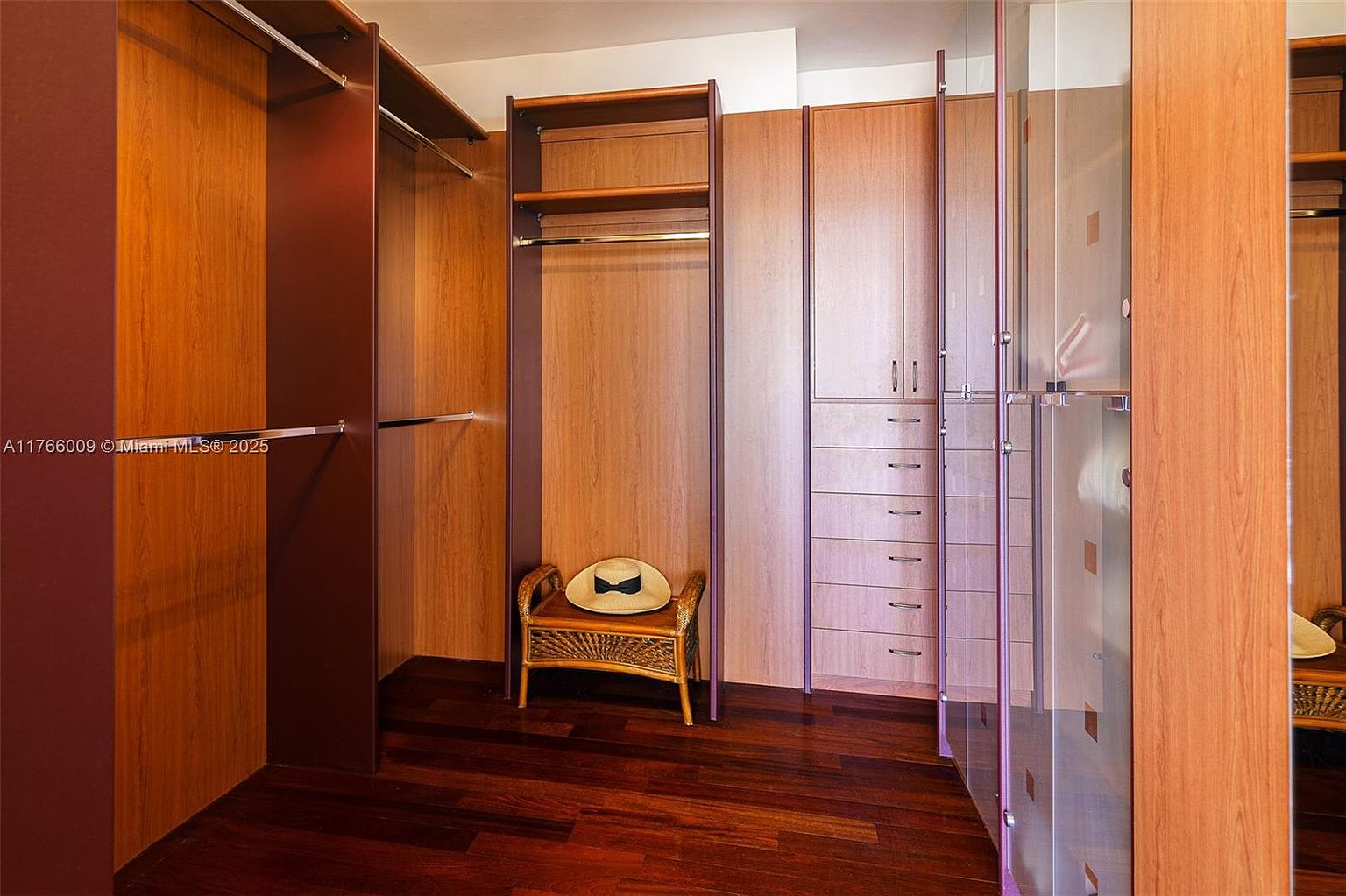 This spacious walk-in closet features warm-toned wood cabinetry with a mix of hanging rods, open shelving, and a built-in dresser unit with multiple drawers. A small, woven-wicker bench with a straw hat sits centrally in the space, resting on rich, dark wood flooring. The perspective is a straight-on view, highlighting the organized storage solutions and the clean, functional layout of the dressing area.