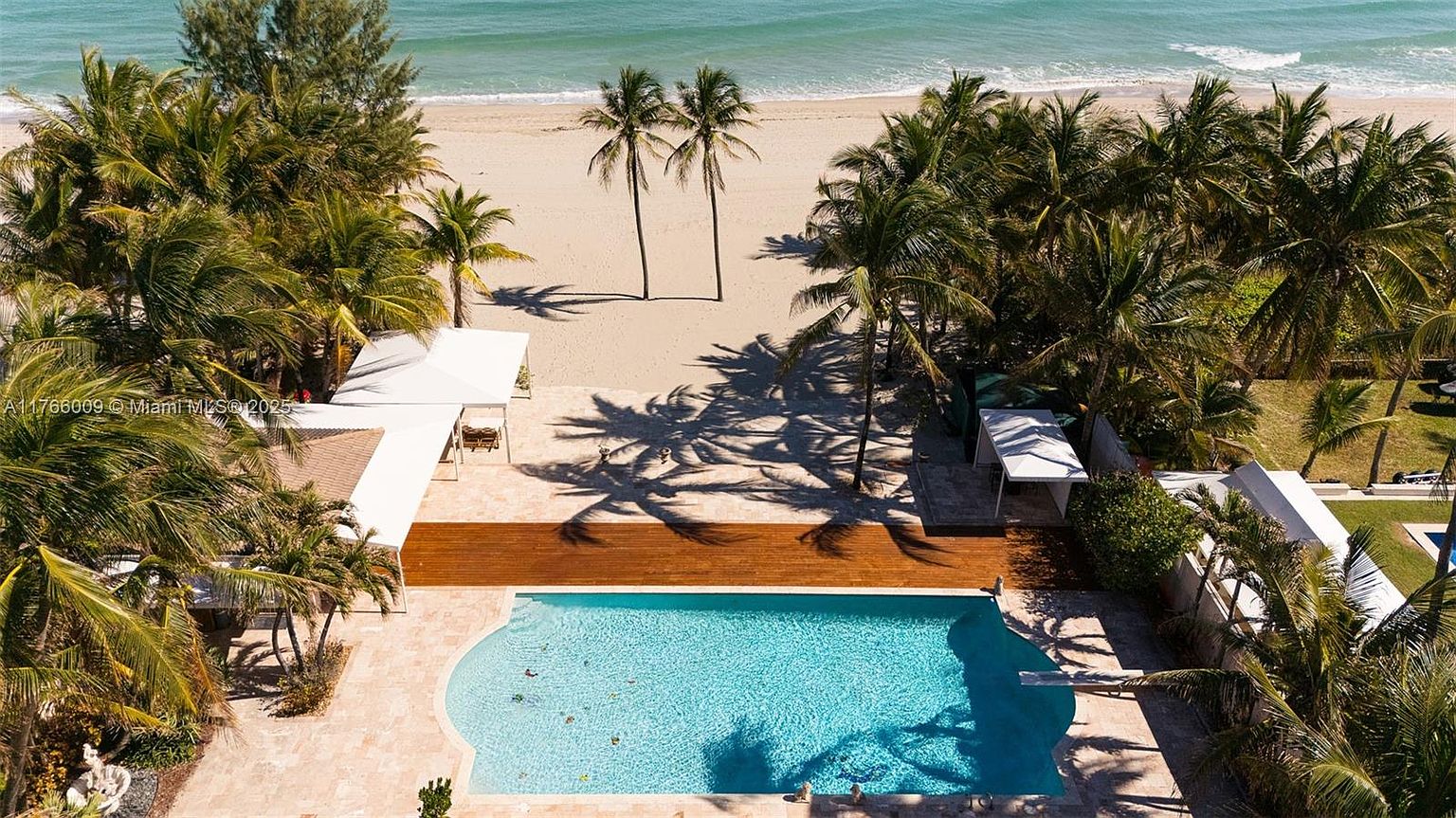 This high-angle aerial shot captures a luxurious beachfront property featuring a large, rectangular swimming pool set within a stone-paved patio. A wooden deck separates the pool area from the sandy beach, which is framed by lush palm trees and direct ocean access. The perspective highlights the seamless transition between the private outdoor living space and the natural coastal environment.