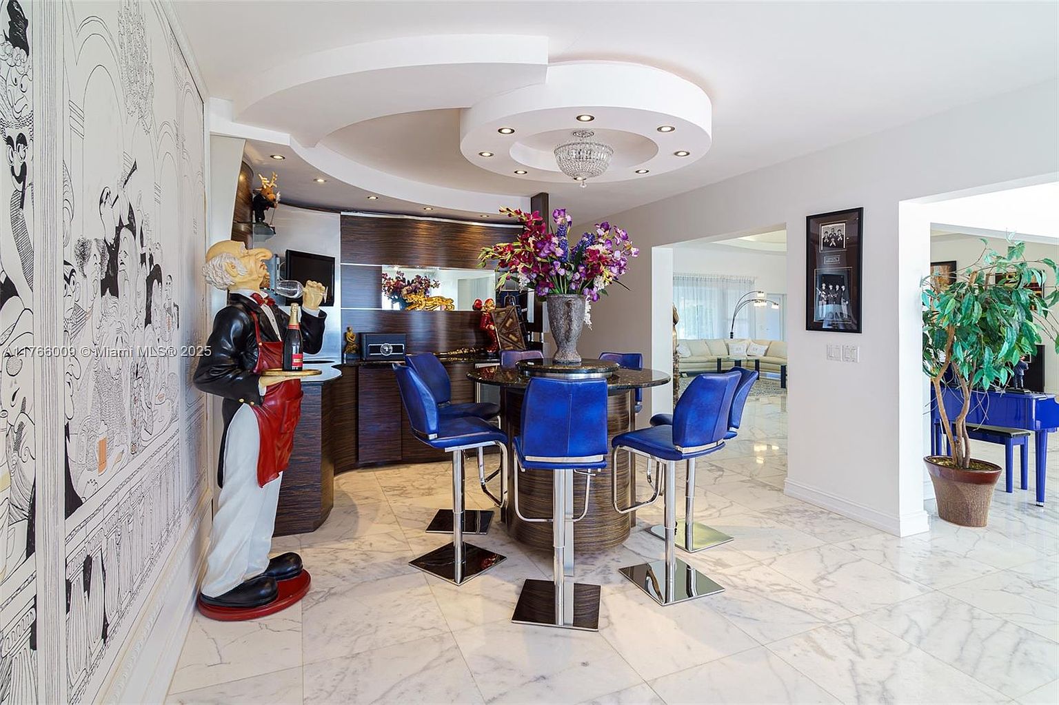 This unique dining area features a circular bar-height table surrounded by four vibrant blue stools, positioned beneath a custom circular ceiling detail with recessed lighting and a central chandelier. To the left, a whimsical butler statue holding a tray adds a playful, eclectic touch to the space, which is finished with polished light-colored stone flooring. The room opens into an adjacent living area, creating an airy, open-concept feel with a view of a blue piano in the background.