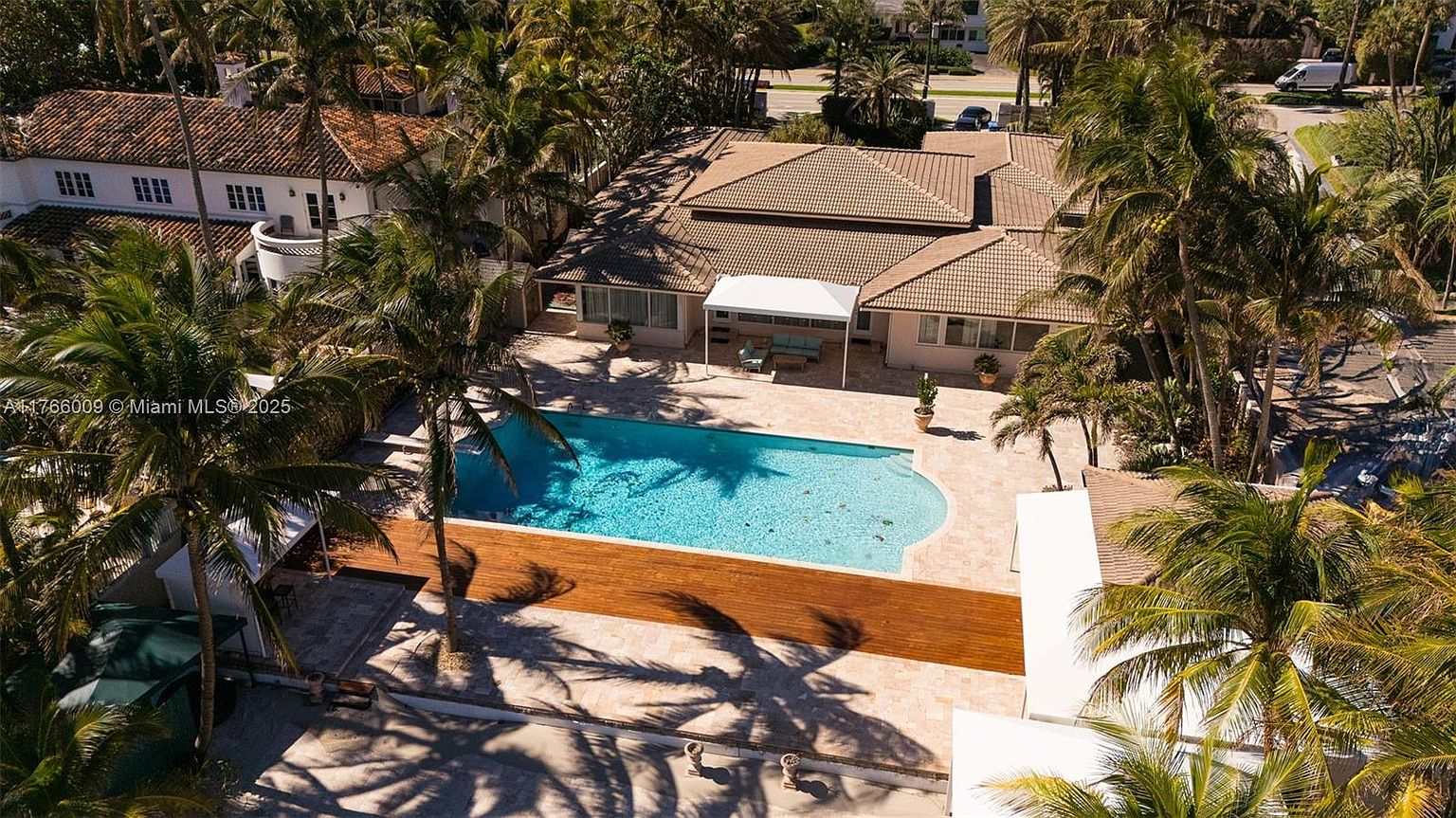 This high-angle aerial view captures a spacious residential property featuring a large, rectangular swimming pool as the central focal point. The backyard includes a paved stone deck, a covered patio area with outdoor seating, and a distinct wooden deck section, all surrounded by lush palm trees. The home itself has a multi-pitched tiled roof and a light-colored exterior, presenting a classic tropical estate aesthetic.