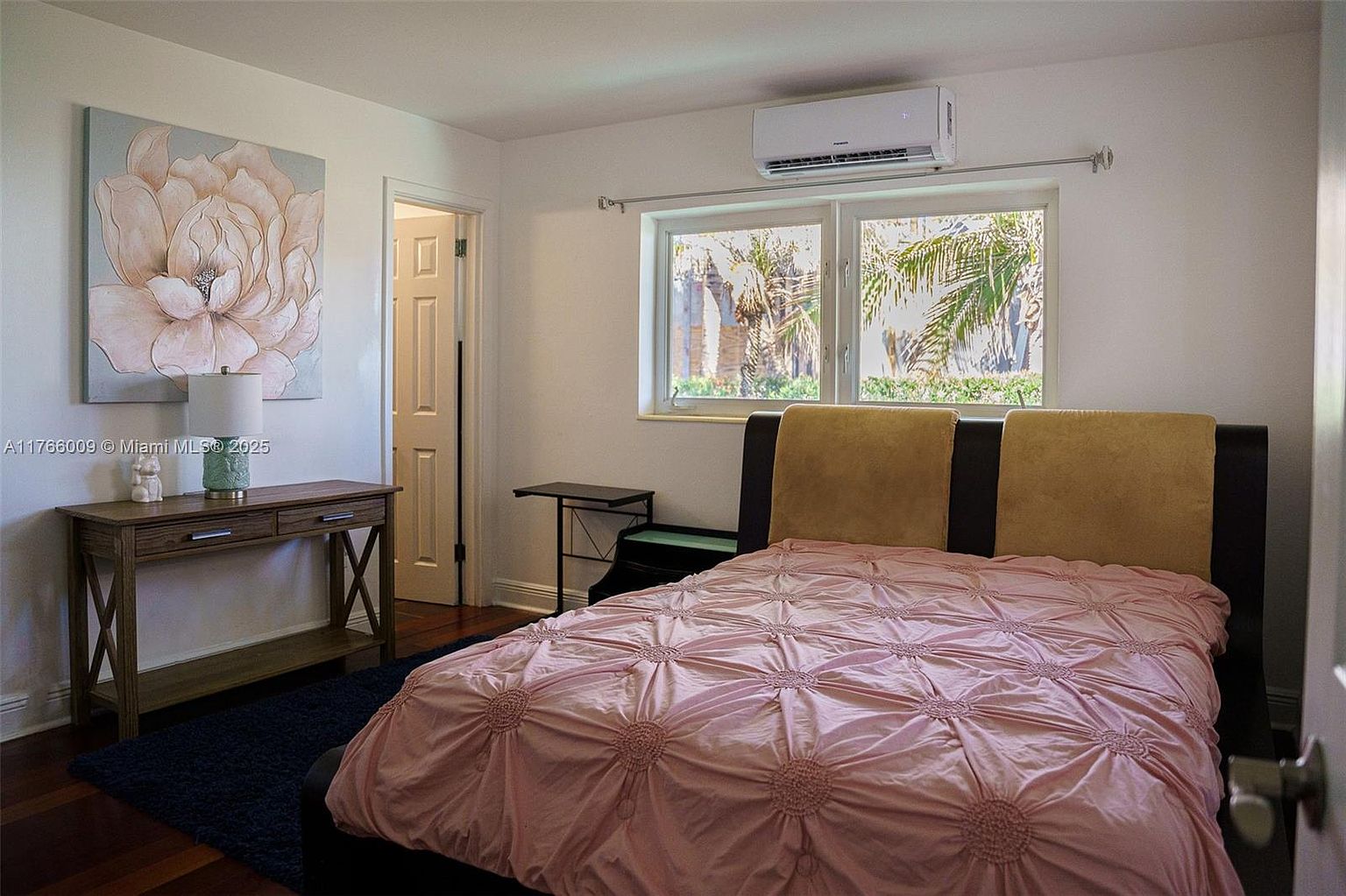 This bedroom features a bed with a tufted headboard and a textured pink comforter, positioned near a window that allows natural light to enter. To the left, a wooden console table holds a lamp and a decorative floral wall art piece, while a doorway leads to an adjacent room. The room is equipped with a wall-mounted air conditioning unit and has dark wood flooring, creating a cozy and functional living space.